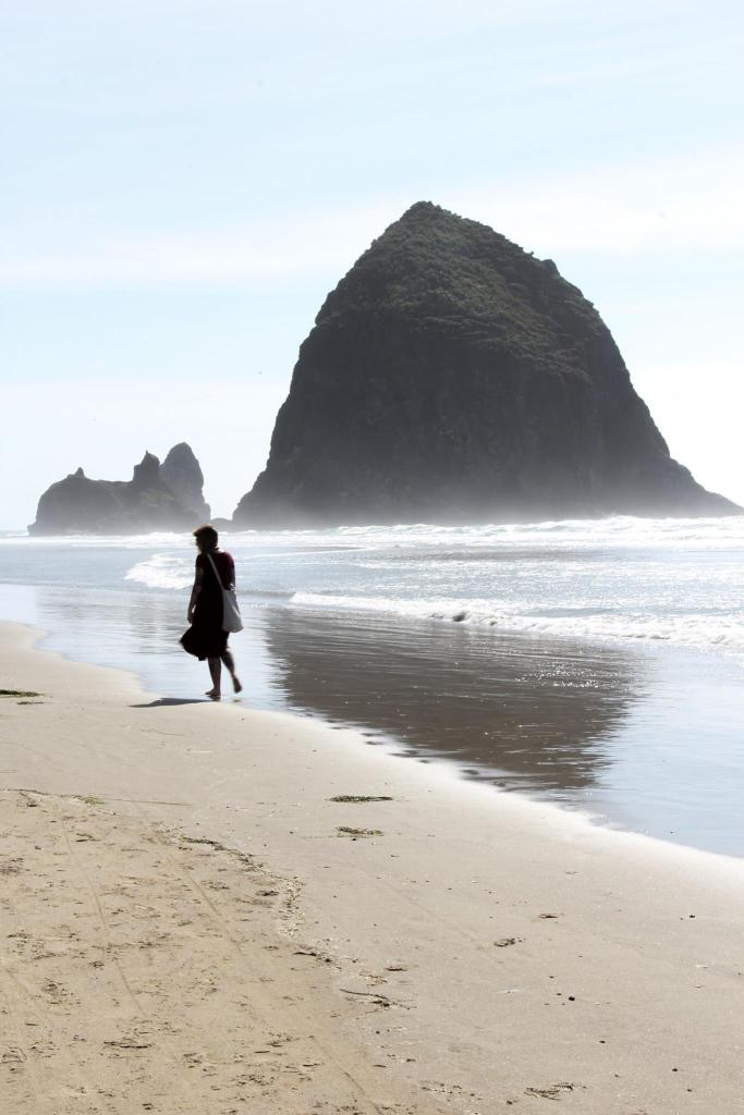 Woman walks along Oregon beach. Giant rock extrudes from the water. 