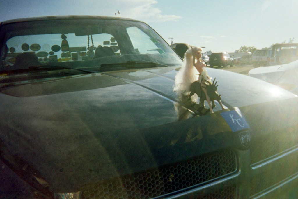Grainy photo of a kid's doll riding a toy triceratops, both placed on the hood of a truck.